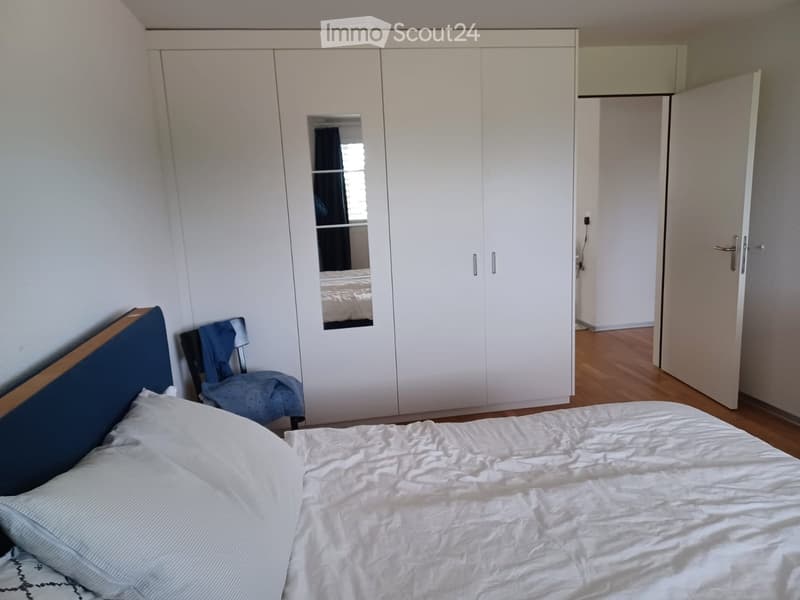 Bedroom with a white wardrobe, a bed with white sheets and pillows, and a blue chair with a folded blanket.