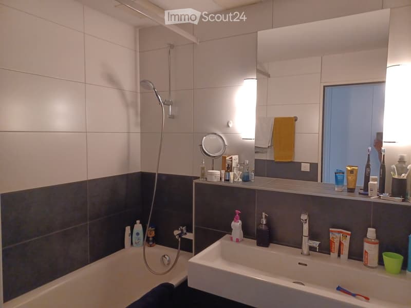 Modern bathroom with a bathtub, showerhead, sink, and various toiletries including toothpaste, soap, and a mirror. The walls are tiled in white and dark gray, and there is a towel hanging on the side.
