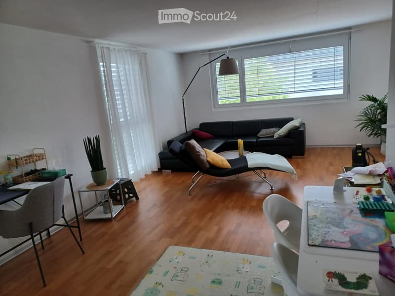 A modern living room with wooden flooring, large windows, a black sectional sofa, a lounge chair, a small table, and a floor lamp.