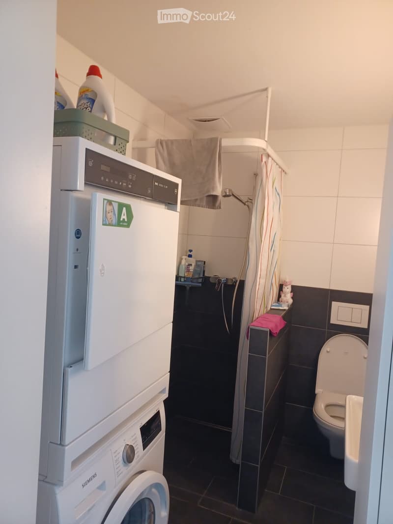 Compact bathroom with a washing machine and dishwasher stacked, a toilet, a shower area with a curtain, and cleaning supplies on a shelf.