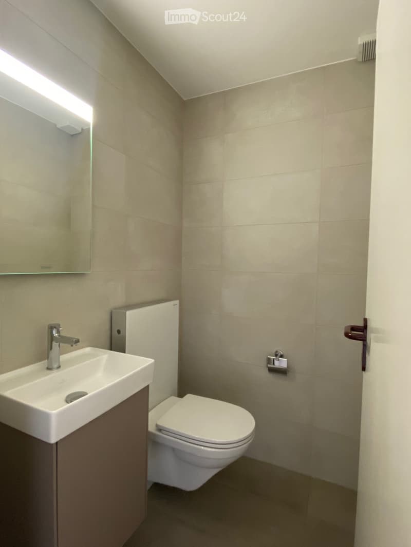 Modern bathroom with a white sink, chrome faucet, toilet, and a mirror above the sink. The walls are tiled in beige, and the floor is dark.