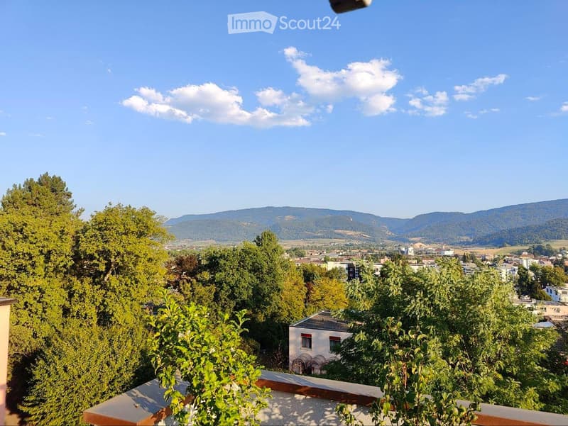 Balcony overlooking a scenic view of a town with trees and distant mountains under a clear blue sky.