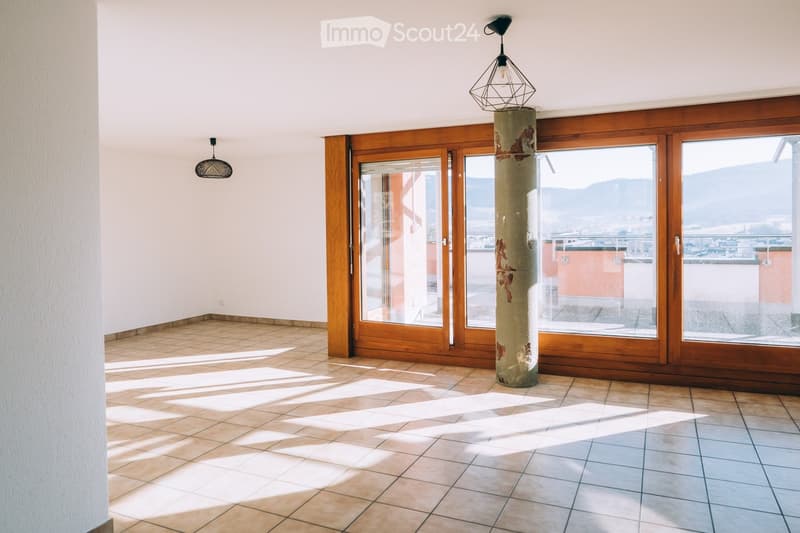 Empty room with tiled floor, large wooden-framed windows, and a central column. Natural light streams in, casting shadows on the floor.