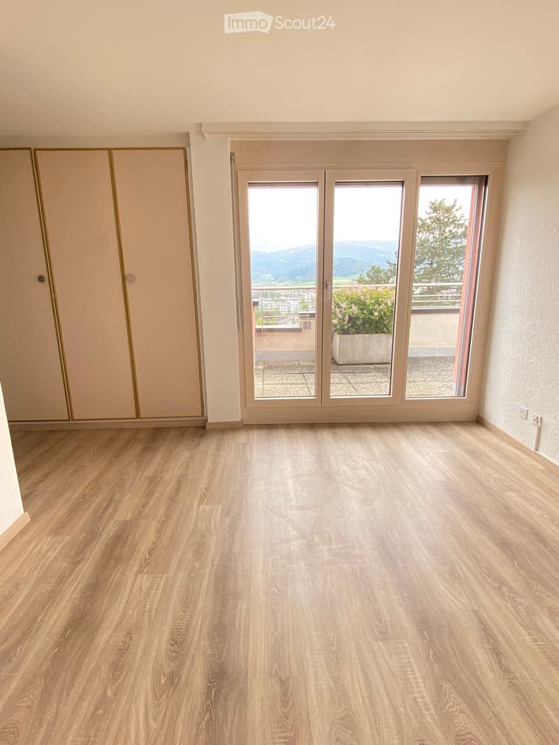 Empty room with light wooden flooring, large sliding glass doors leading to a balcony with a view of mountains and trees.