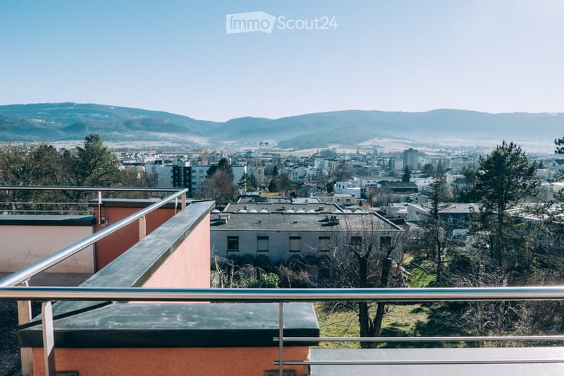 Balcony with metal railing overlooking a cityscape, with buildings, trees, and distant hills visible.