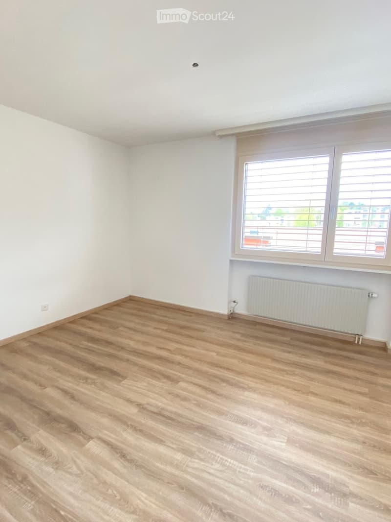 Empty room with light wooden flooring, white walls, and a large window with blinds. A radiator is visible below the window.