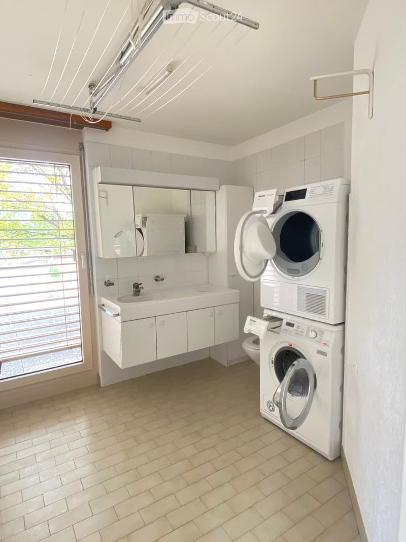 Bright bathroom with a white sink, mirror, and stacked washing machine and dryer unit. The space features tiled walls and a window with blinds.