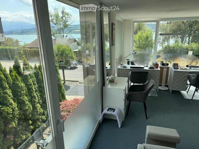 Modern office space with large windows overlooking a scenic lake and mountains, featuring a desk, chairs, a small table, and a stool.
