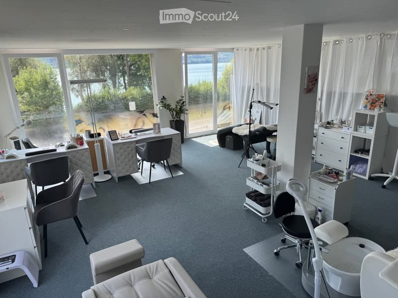 Modern, well-lit salon with large windows, grey carpet, white furniture, and a view of the water.