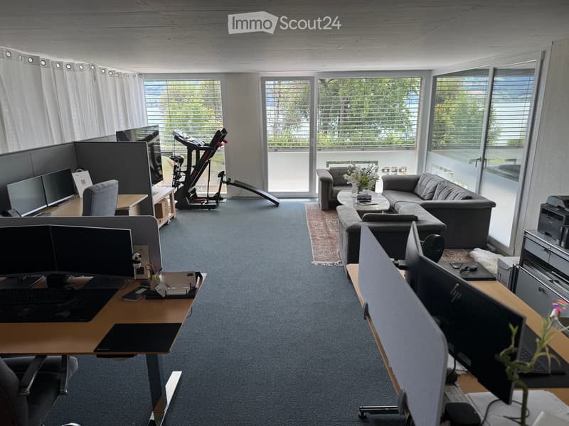 Office space with desks, chairs, laptops, a sofa, coffee table, and a fitness bike. Large windows with blinds and a view of greenery.