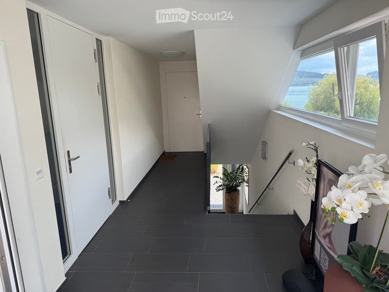 Modern hallway with dark tiled flooring, white walls, large windows offering a scenic view, and a staircase leading upwards.