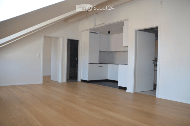 Empty room with wooden flooring, white walls, and a small kitchen area with white cabinets and a sink.