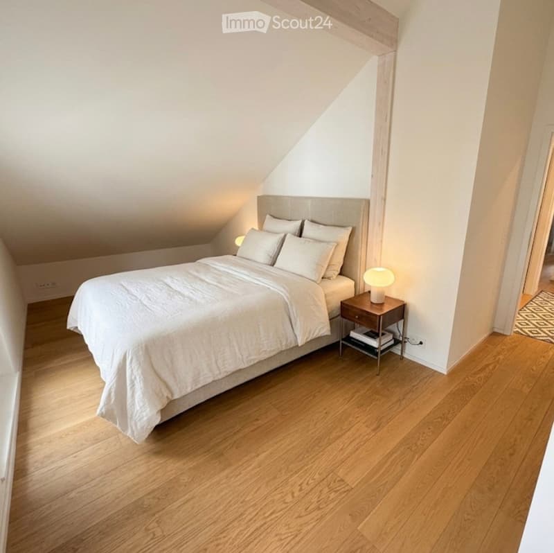 Cozy attic bedroom with wooden flooring, a large bed with white bedding, a nightstand with a lamp, and books on the nightstand.