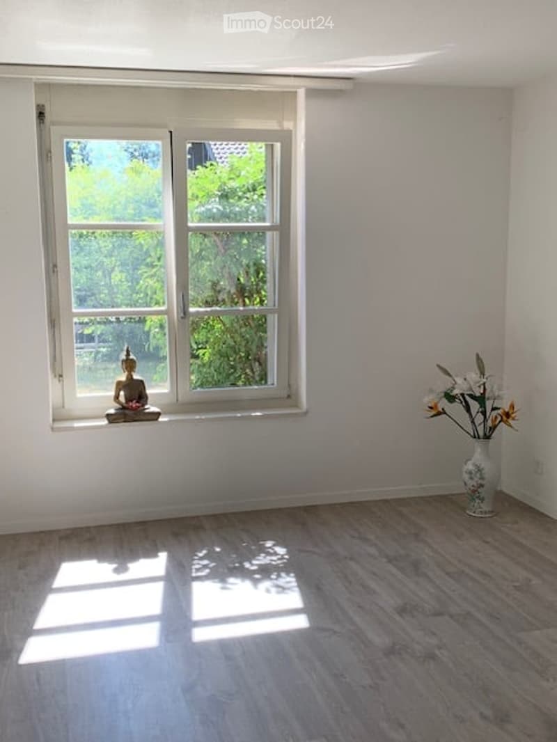 Empty room with light wooden flooring, white walls, and a large window showing greenery outside. A small decorative item sits on the windowsill, and a vase with flowers is placed in the corner.
