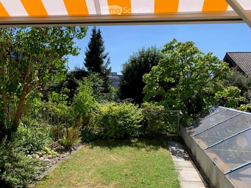 Well-maintained garden with a lawn, various trees, shrubs, and a small greenhouse. The area is enclosed and features a clear, sunny sky.