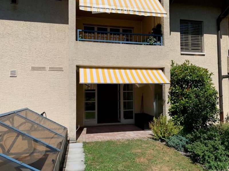 Two-story building with beige walls, blue railings, striped awnings, and a small garden area.