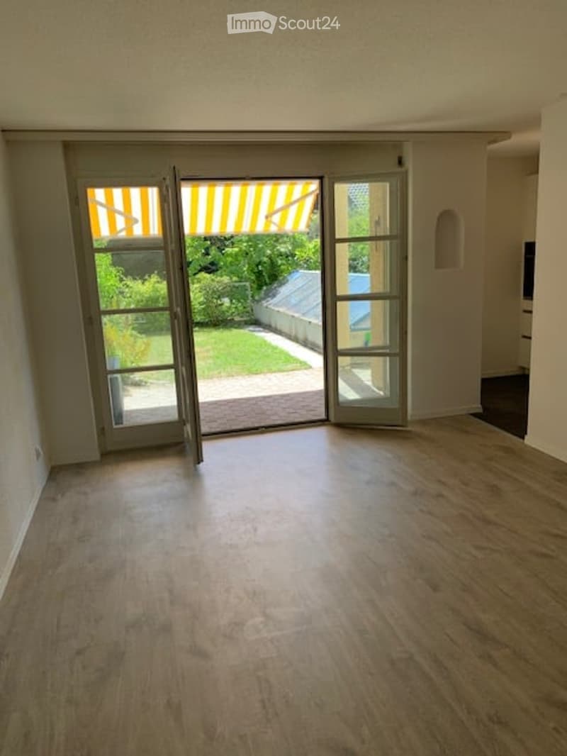 Empty room with wooden flooring, large sliding glass doors leading to a patio with a striped awning and a view of a garden.