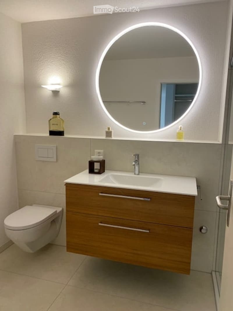 Modern bathroom with a wooden vanity, round LED-lit mirror, sink, and toilet. The vanity has two drawers and a sleek faucet. A small shelf holds toiletries and a decorative bottle.