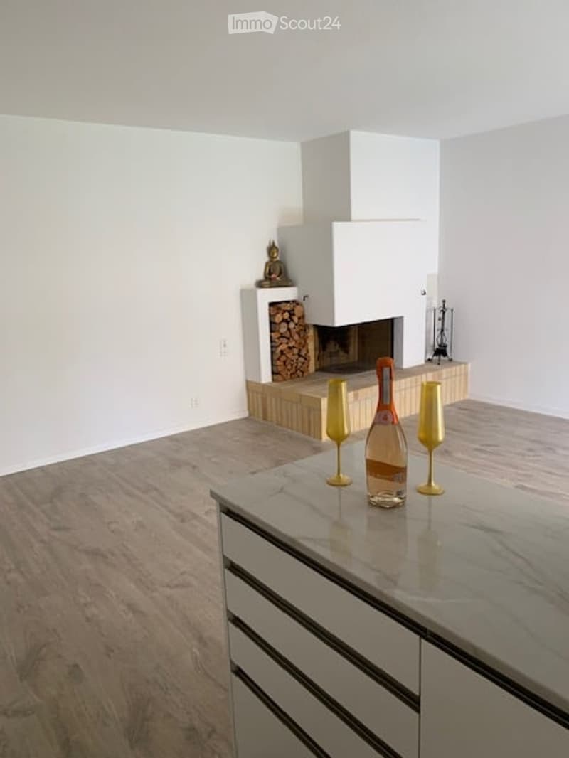 Modern living room with a white fireplace, marble floor, and a small bar area with wine glasses and a bottle. A decorative statue sits near the fireplace.