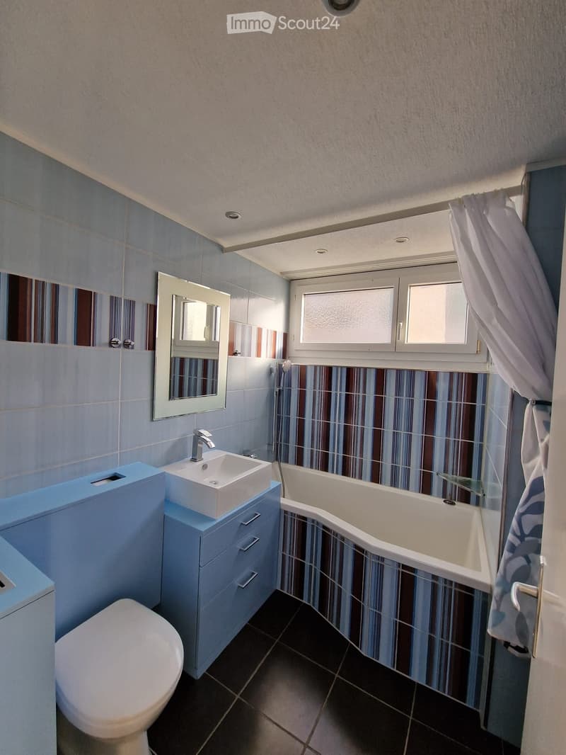 Modern bathroom with a white sink, blue cabinet, bathtub, toilet, and a large mirror above the sink. The walls are tiled in blue and brown stripes, and there is a window with a curtain.
