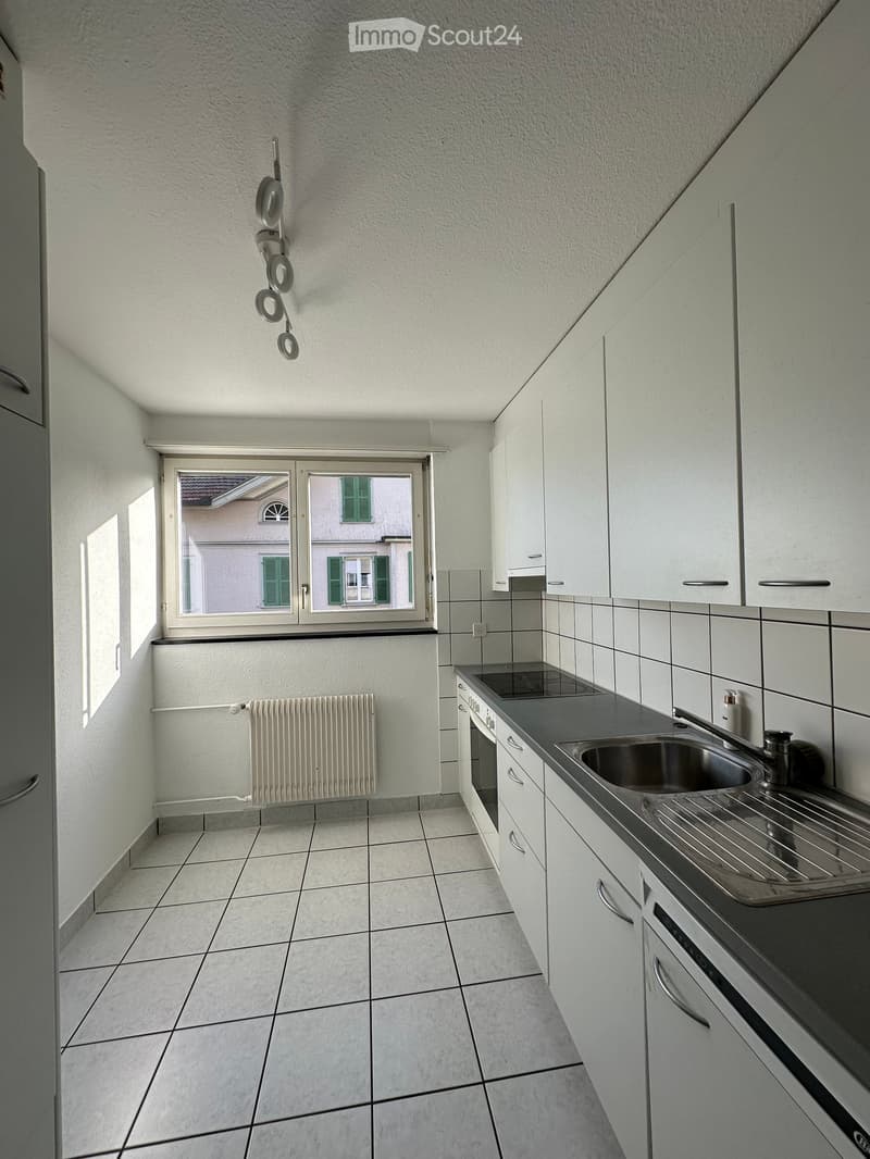 Modern kitchen with white cabinets, stainless steel sink, oven, and radiator. The space features tiled flooring and a window offering a view of the street.