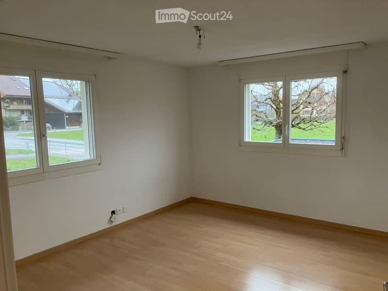 Empty room with light wooden flooring, white walls, and large windows offering a view of the street and neighboring houses.