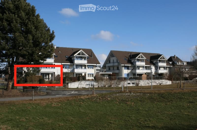 Multi-story residential building with white walls, brown roof, and balconies. The building is surrounded by a grassy area and trees.