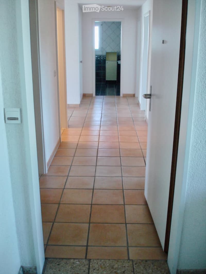 A long, narrow hallway with tiled flooring and white walls, leading to an open doorway at the end.