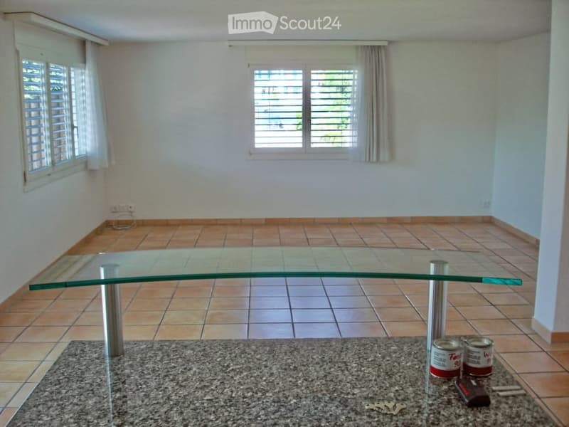 Empty room with tiled floor, large glass table, and a few items on the floor including cans, keys, and a remote control.