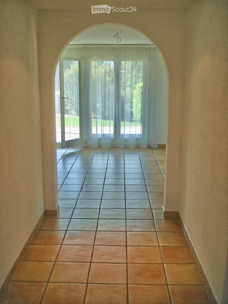 A tiled hallway with an arched doorway leading to a room with large windows and sheer curtains. The floor is tiled in two different colors, and the walls are painted a light beige.