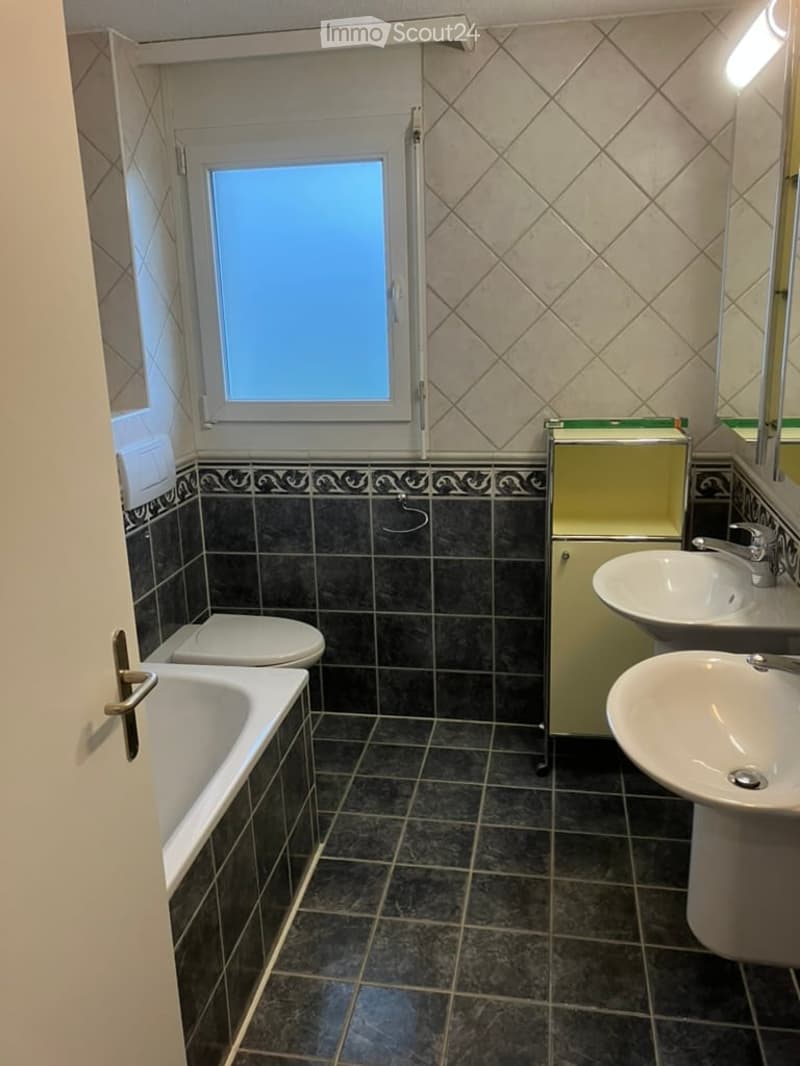 Bathroom with dark tiled walls and floor, white bathtub, toilet, two sinks, and a mirrored cabinet. A small window provides natural light.
