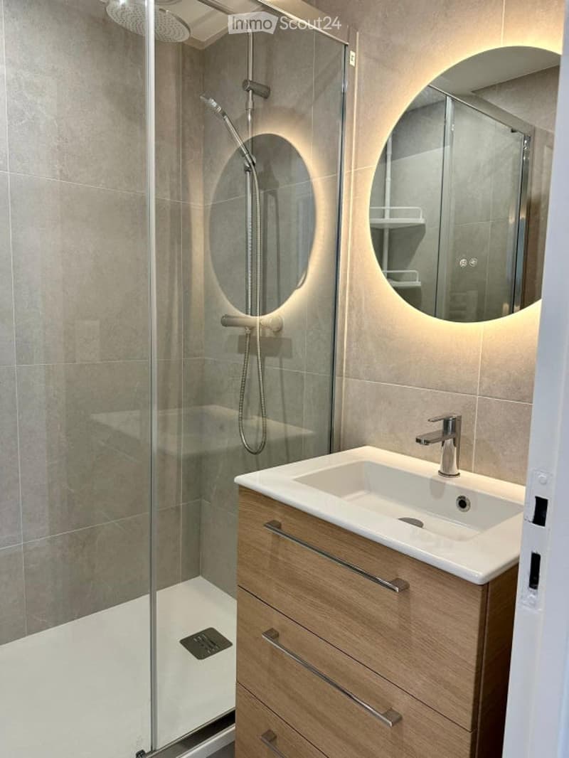 Modern bathroom with a glass-enclosed shower, a white sink with a wooden cabinet, and a round mirror with built-in lighting.