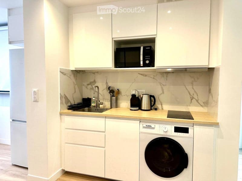 Modern kitchen with white cabinets, marble backsplash, and wooden countertops. Features include a sink, microwave, refrigerator, washing machine, and electric stove.