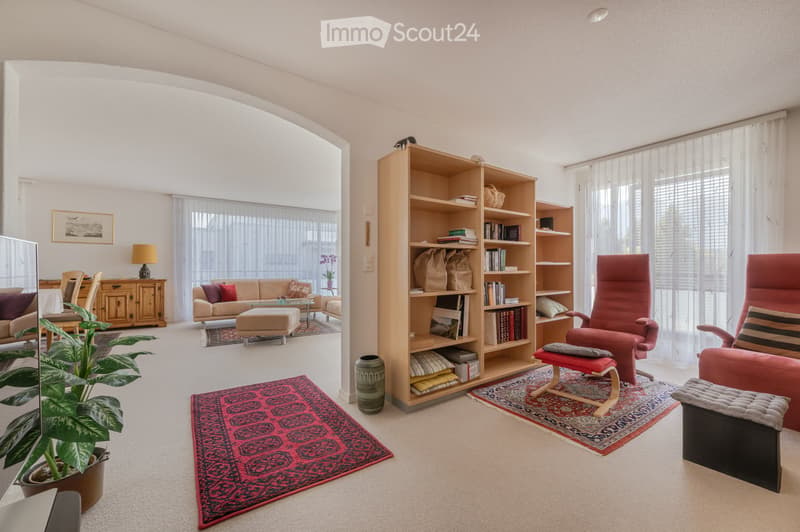 Bright living room with large windows, cream-colored sofa, red accent chairs, wooden bookshelf, and decorative rugs.