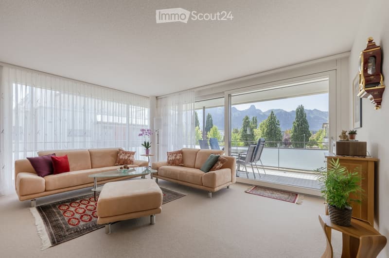 Bright living room with large windows and a balcony overlooking a scenic mountain view. Features a beige sofa set, glass coffee table, ottoman, and decorative pillows.