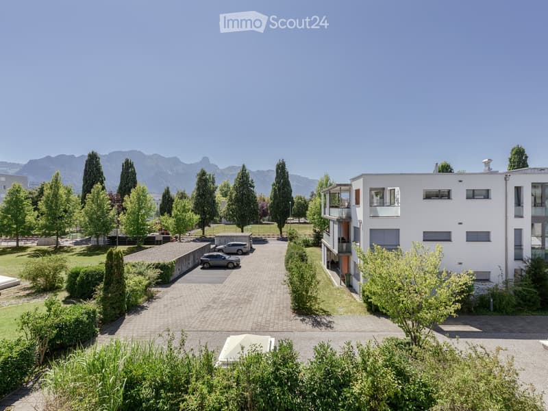 Modern multi-story building with balconies, surrounded by landscaped gardens, trees, and parked cars. The background features a scenic mountain view.