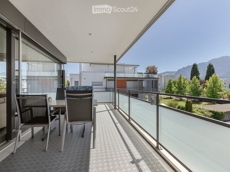 Modern balcony with a glass railing, a small table, and four chairs, overlooking a landscaped area with trees and mountains in the background.