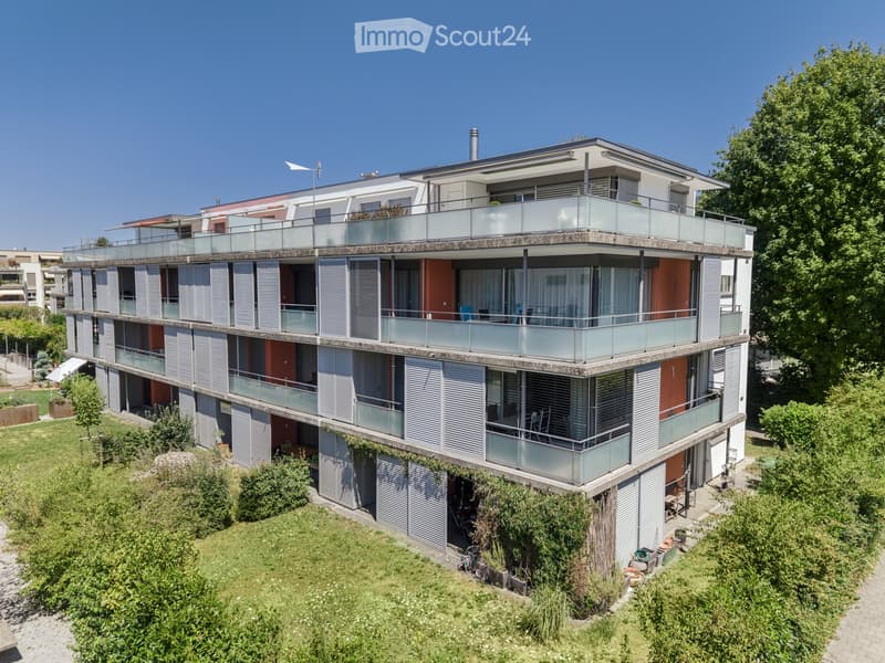 Modern multi-story residential building with balconies, glass railings, and contemporary design, surrounded by greenery.