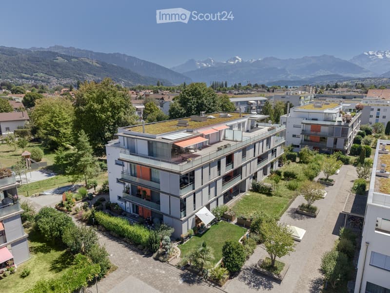 A modern multi-story residential building with balconies, surrounded by greenery and a scenic mountain backdrop.