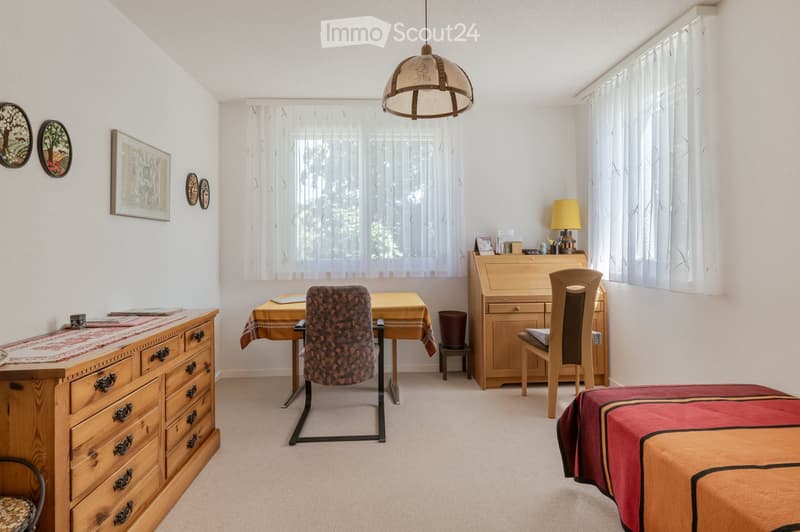 A well-furnished room with a bed, chairs, a table, a lamp, a cabinet, and picture frames on the wall.
