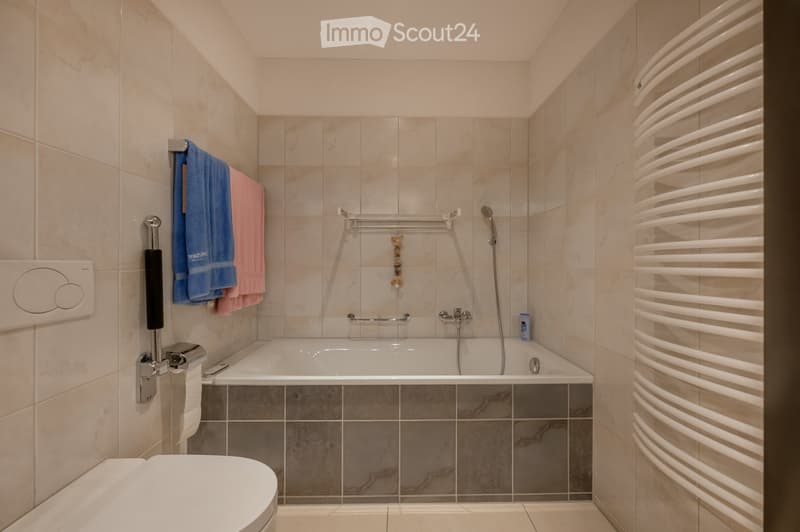 Modern bathroom with a bathtub, toilet, towel rack with towels, showerhead, and a radiator. The walls are tiled with beige tiles, and the floor has a marble pattern.