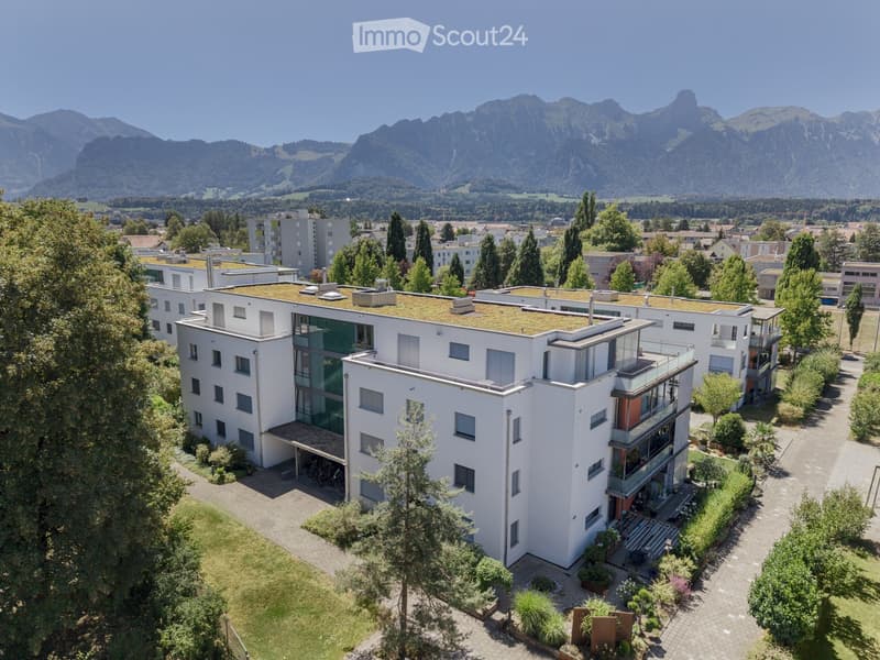 Modern white multi-story building with a green roof, surrounded by trees and greenery, with a scenic mountain backdrop.