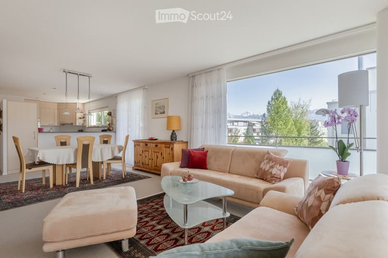 Bright living and dining area with large windows offering a scenic view, featuring a beige sofa, glass coffee table, dining table with chairs, and a wooden cabinet.