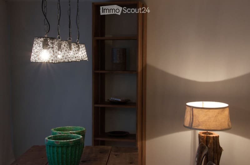 A cozy, well-lit room with a wooden table holding two green vases, a wooden shelf with decorative items, and a table lamp with a beige shade on a wooden base.