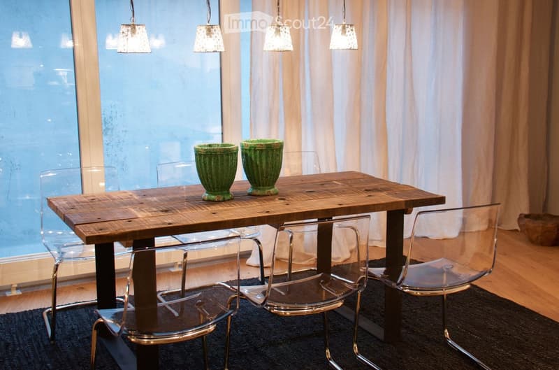 A modern dining area with a wooden table, four transparent chairs, and two green vases on the table. The room features large windows with sheer curtains and pendant lights hanging from the ceiling.