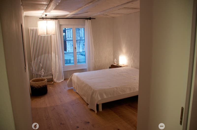 Cozy bedroom with wooden flooring, a double bed with white bedding, a nightstand with a lamp, a wicker basket, a metal chair, and a radiator near the window with white curtains.