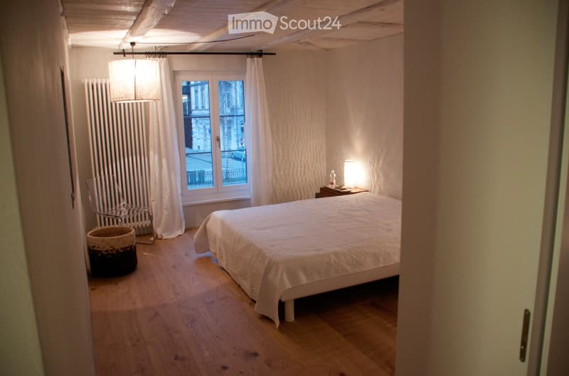 Cozy bedroom with wooden flooring, a double bed with white bedding, a nightstand with a lamp, a wicker basket, a metal chair, and a radiator near the window with white curtains.