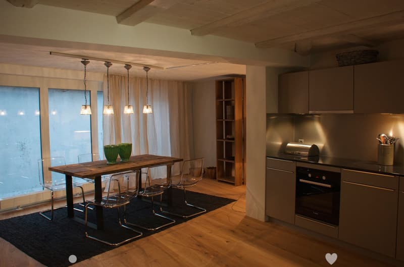 Modern open-plan kitchen and dining area with wooden flooring, pendant lights, a wooden dining table with four transparent chairs, and a grey kitchen with built-in appliances.