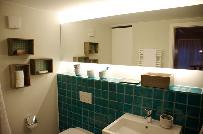 Modern bathroom with teal tiles, a white sink, a toilet, a large mirror, and wooden shelves holding toiletries and towels.
