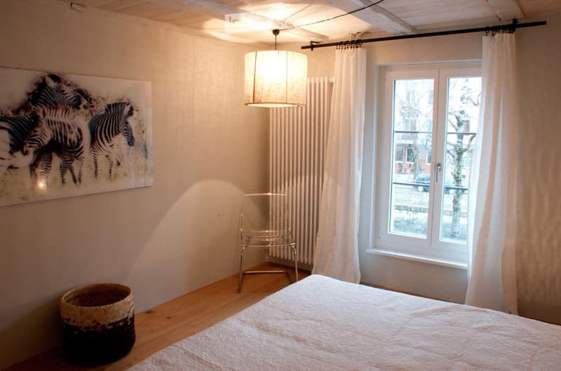 A modern bedroom with a large bed, a wicker basket, a metal chair, a large picture frame of zebras, and a hanging lamp. The room has wooden flooring and large windows with white curtains.